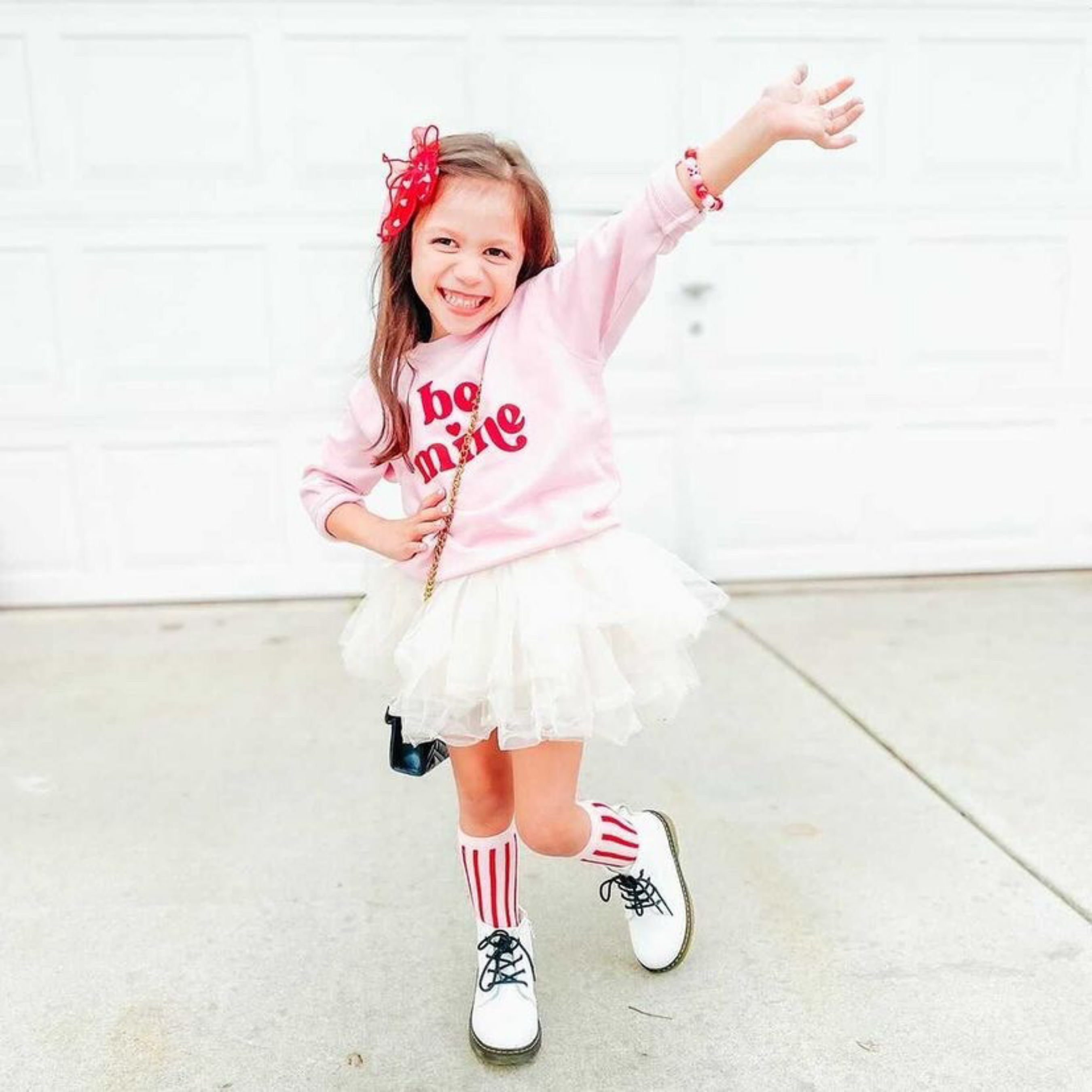 Child wearing a pink 'be mine' sweatshirt with a white tutu and striped socks, standing on a light-colored floor.