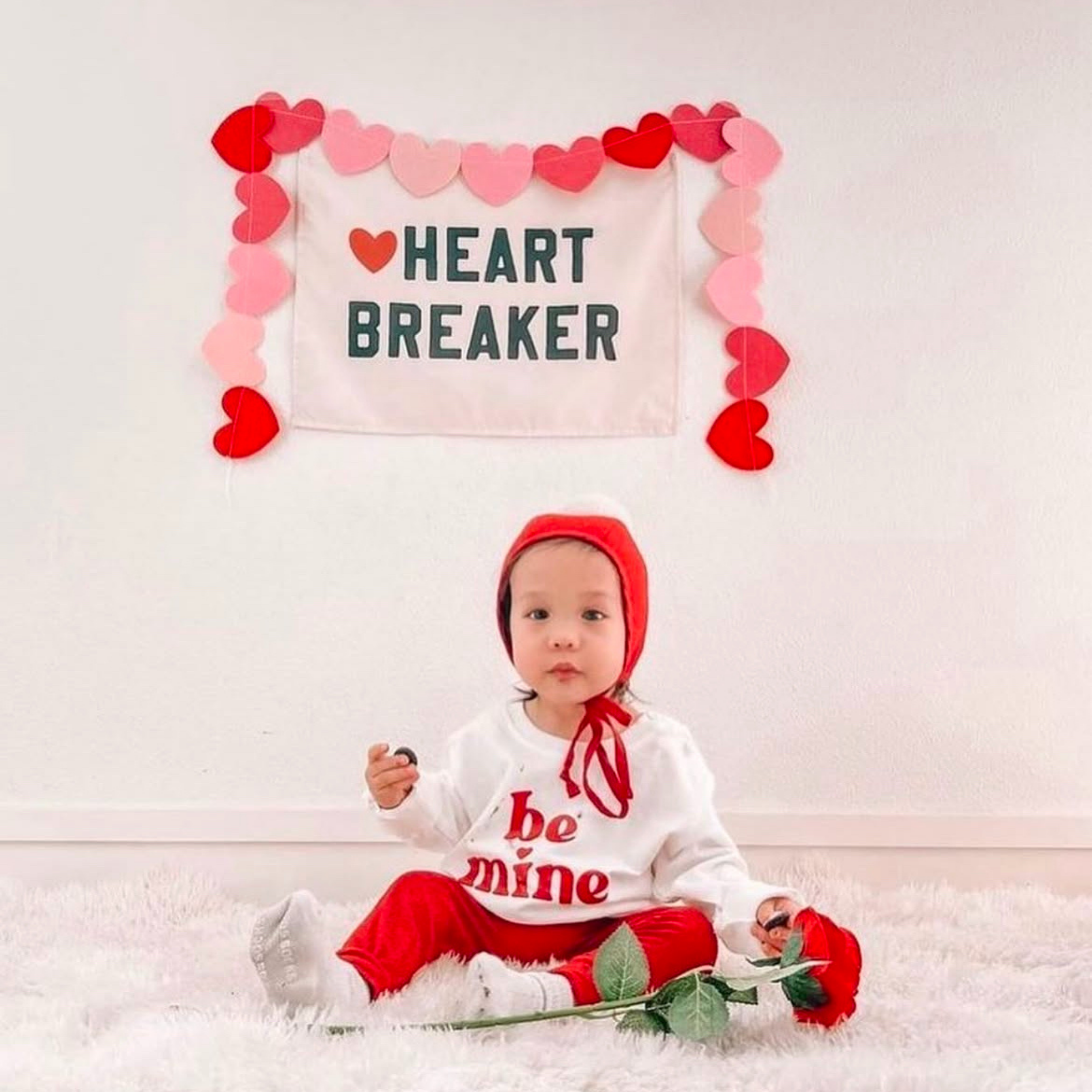 Baby in Valentine's Day outfit with 'be mine' shirt and red heartbreaker sign.