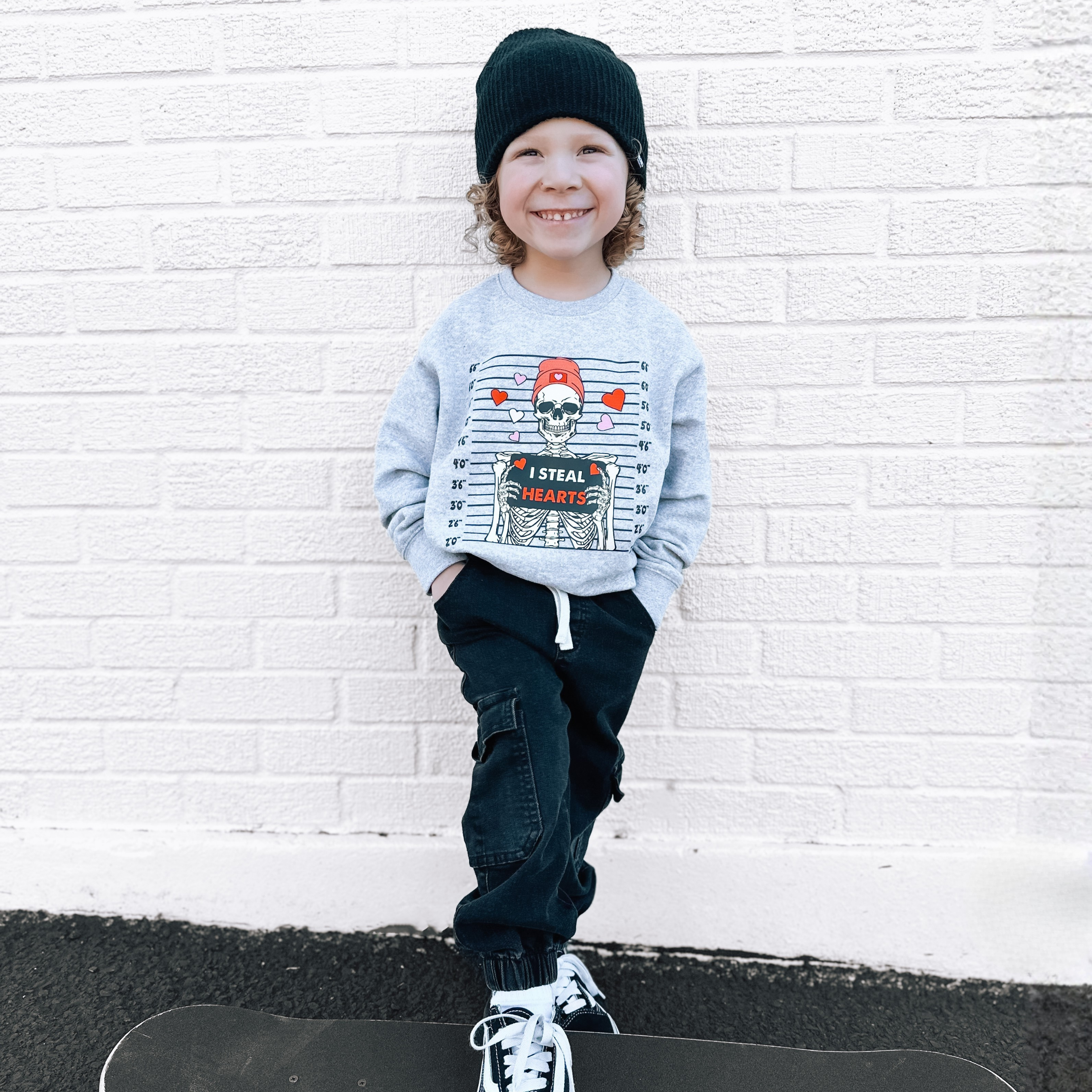 Child wearing a gray sweatshirt with a skeleton Valentine's graphic design, black pants, and a black beanie, sitting on a skateboard against a white brick wall.