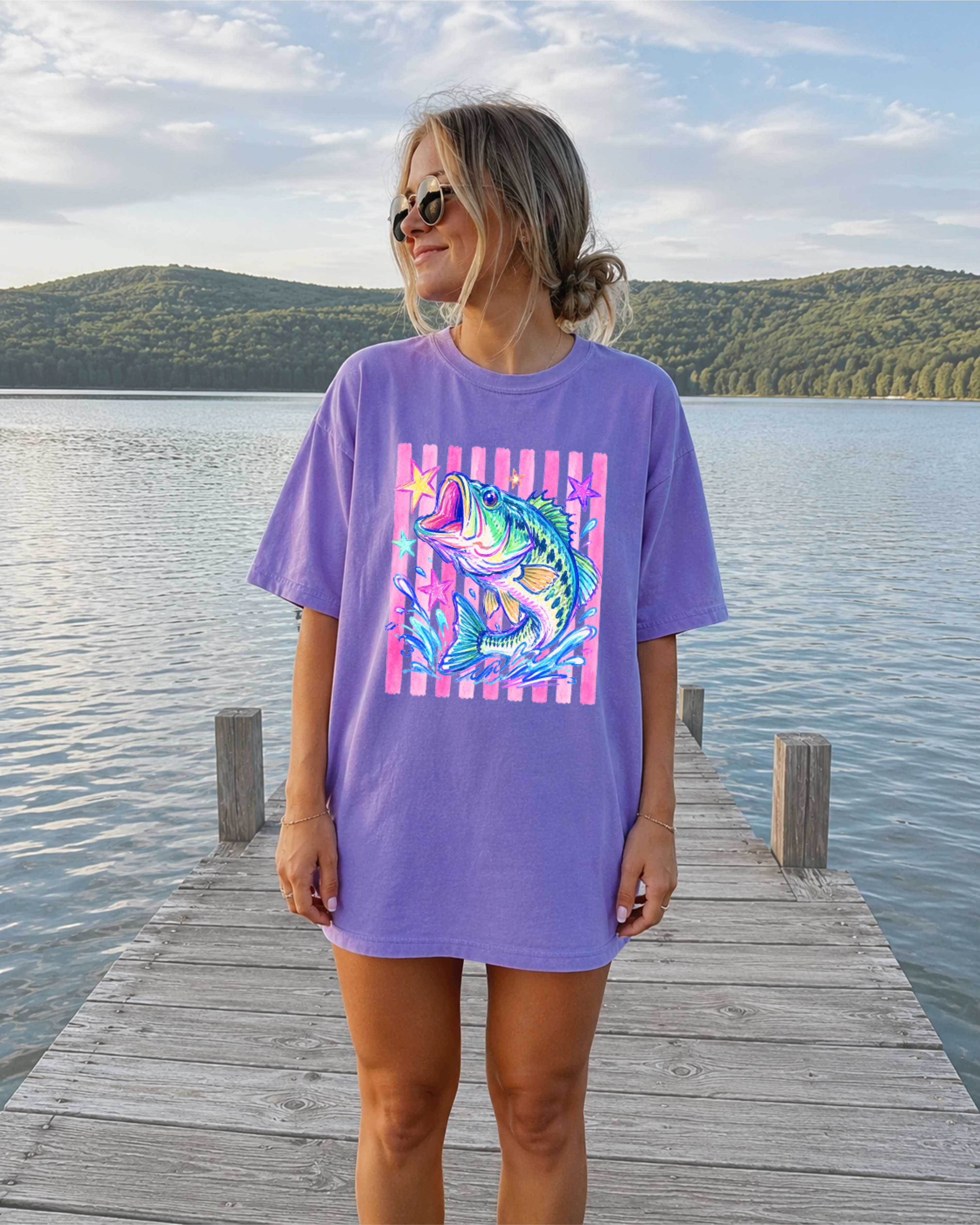 Woman wearing a violet Comfort Colors t-shirt with colorful illustrated bass fish graphic, standing on a dock by a lake at sunset