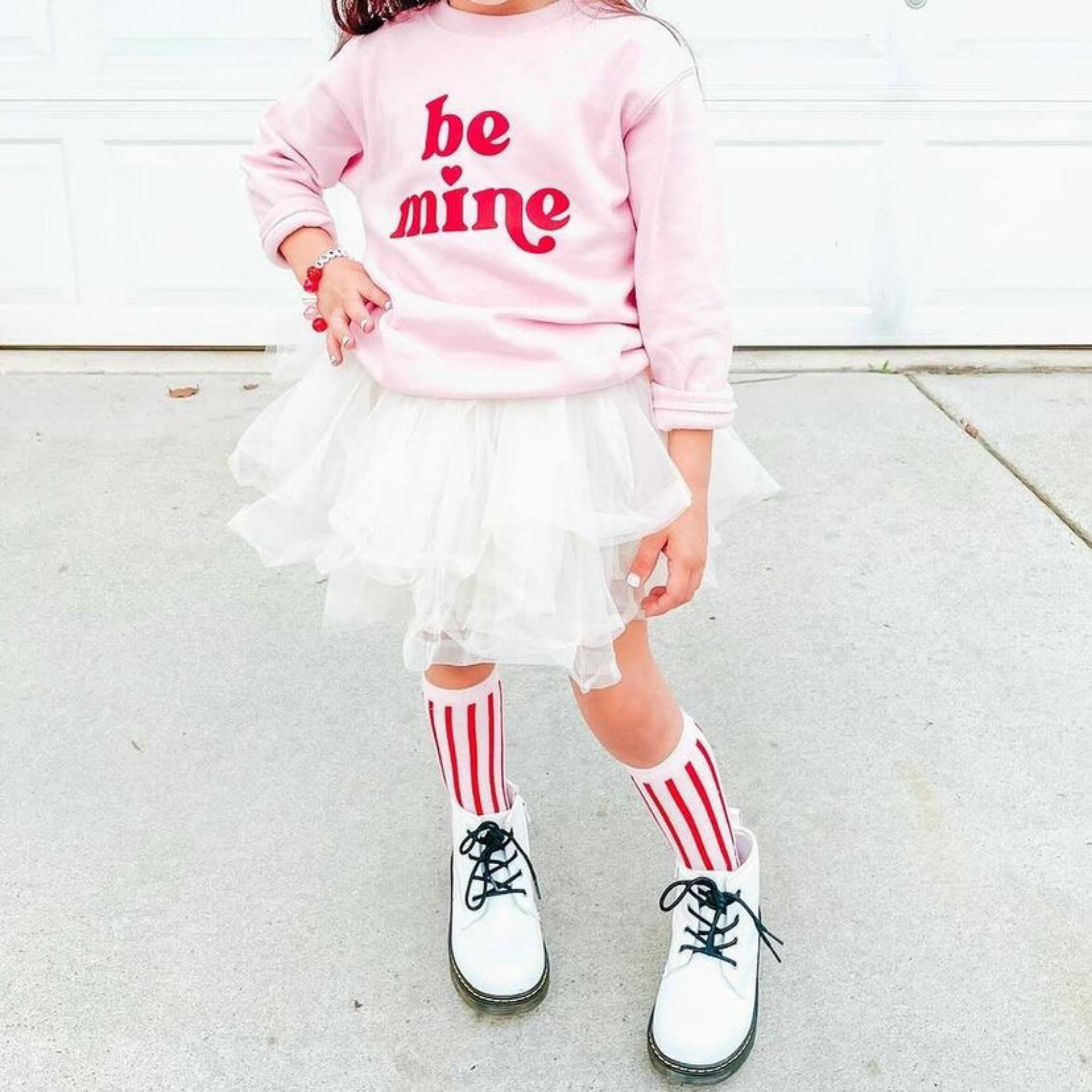 Child wearing a pink 'be mine' sweatshirt, white skirt, red and pink striped socks, and white shoes on a light background.