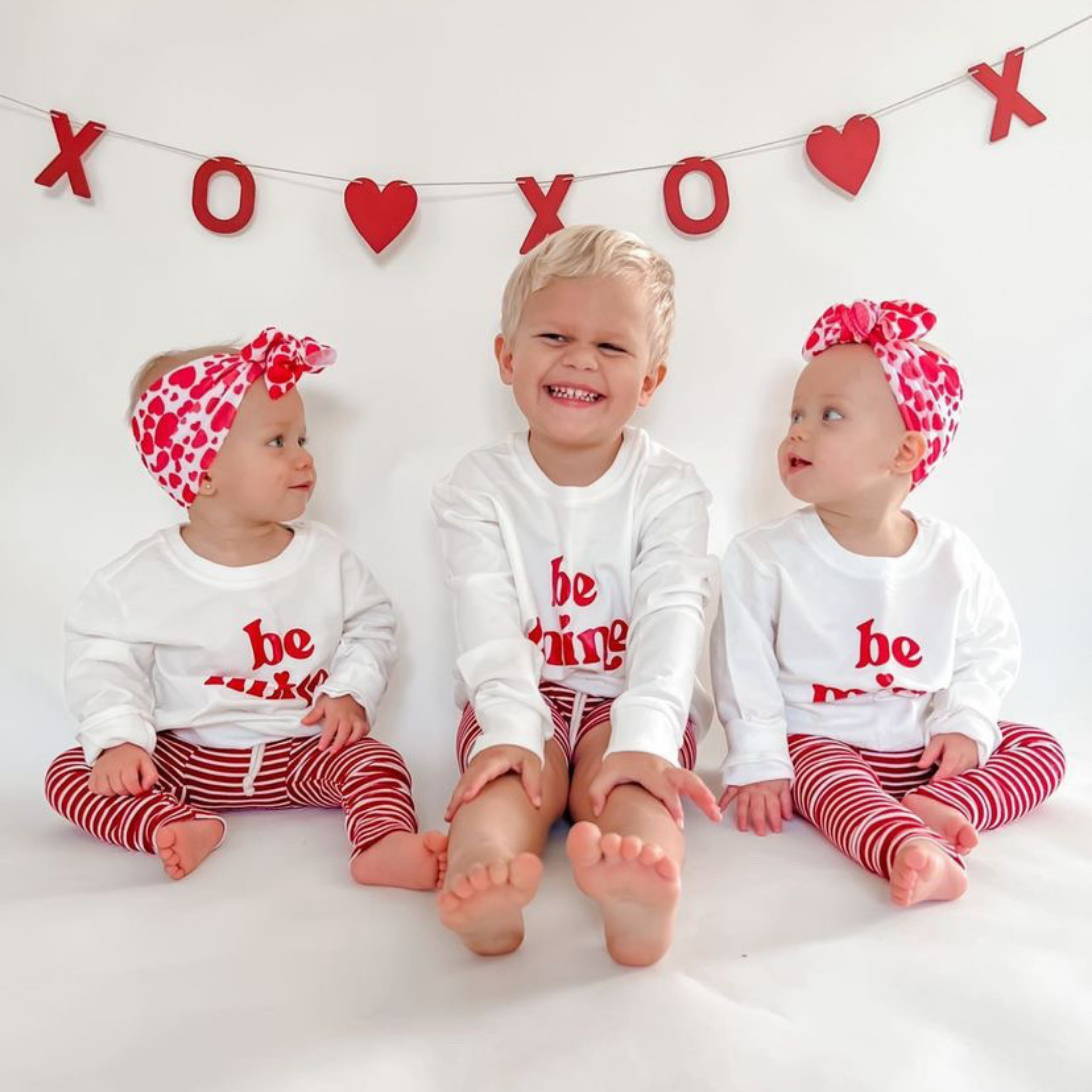Three children wearing matching Valentine's Day outfits with a decorative banner in the background.