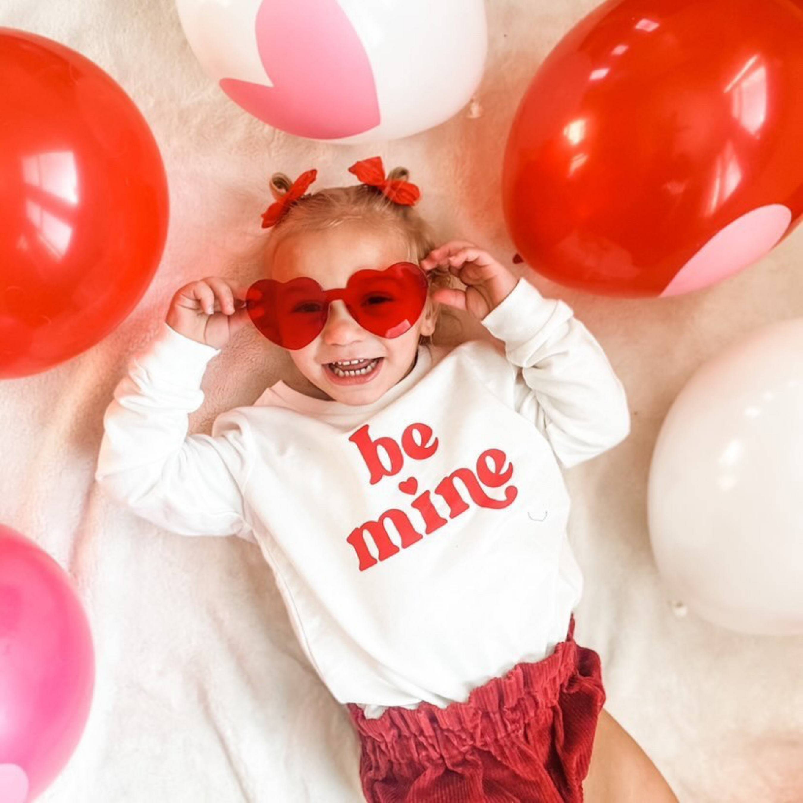 Child wearing a 'be mine' shirt surrounded by balloons