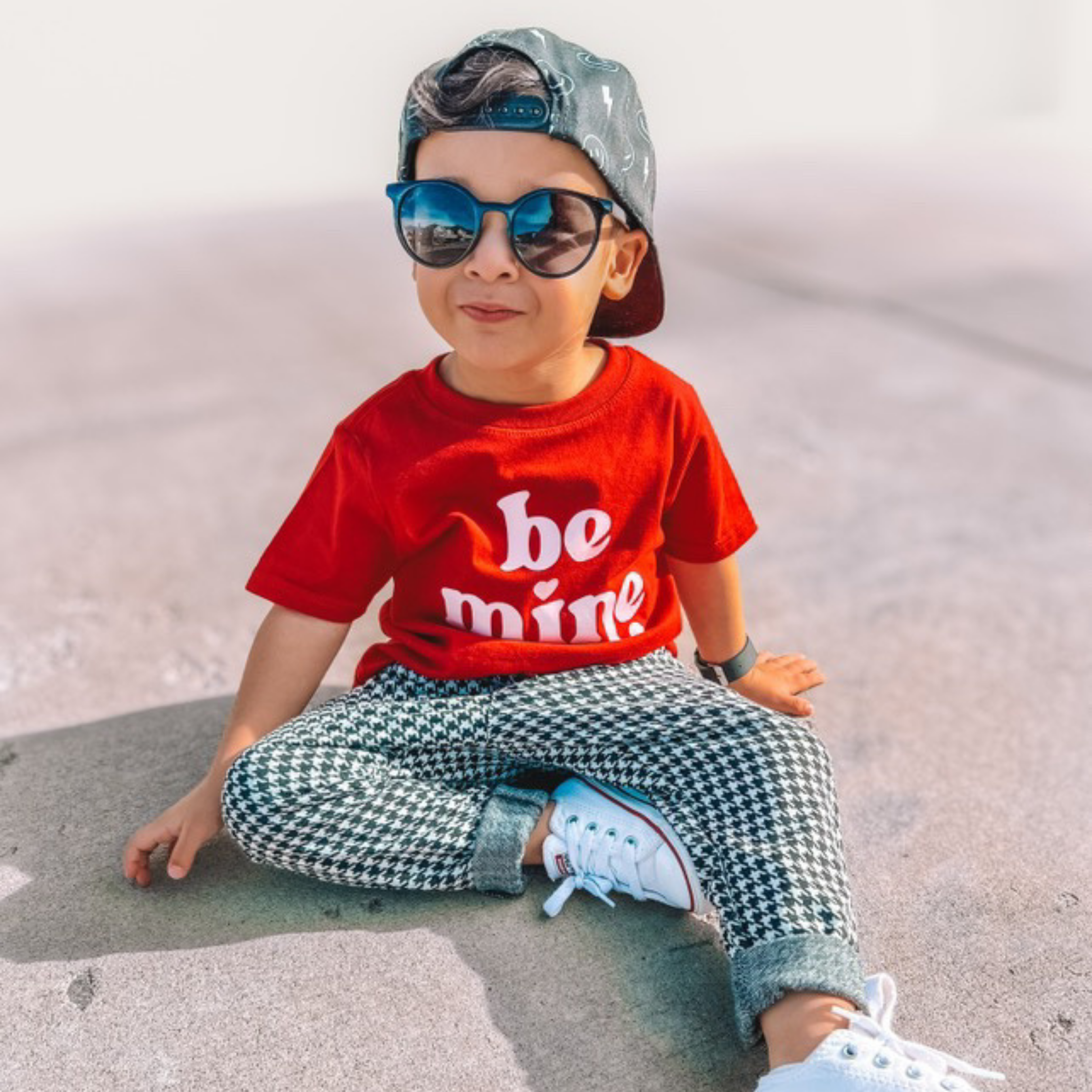 Child wearing a red shirt with 'be mine' text, sunglasses, and checkered pants sitting on a concrete surface.