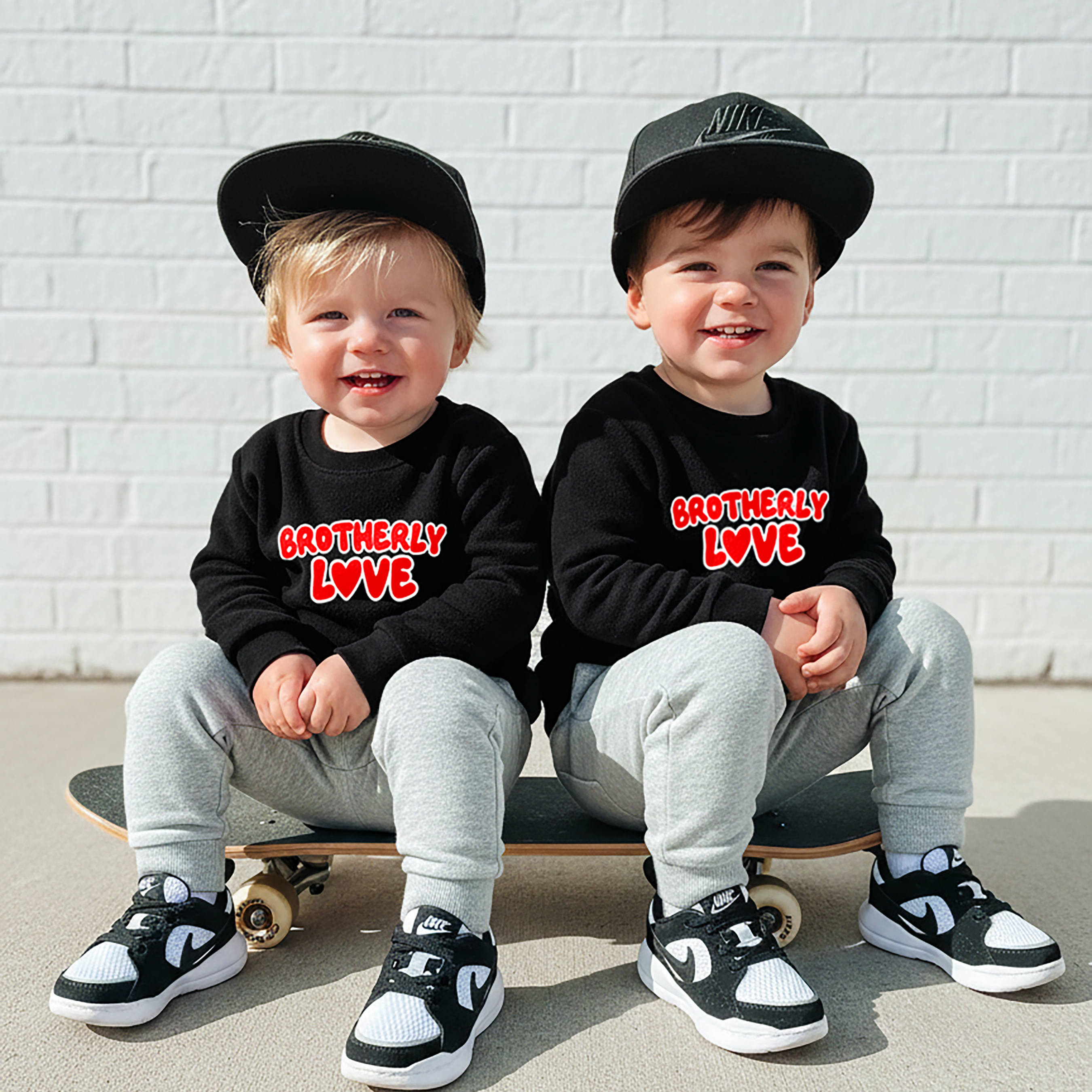 Two children sitting on skateboards wearing matching black sweatshirts with 'Brotherly Love' text, gray pants, and black caps.