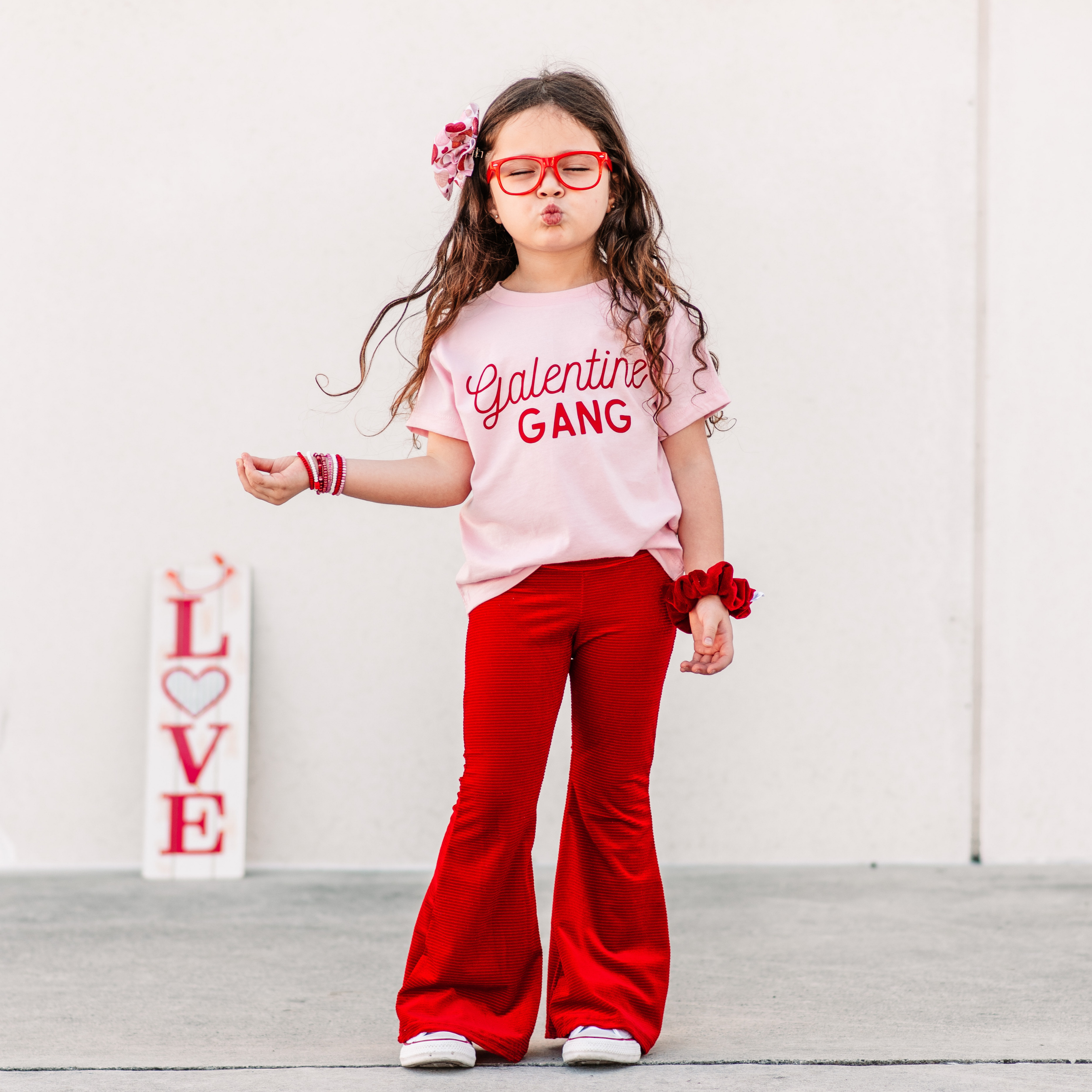 Child wearing a pink shirt with 'Galentine Gang' text and red pants, posing against a white background.