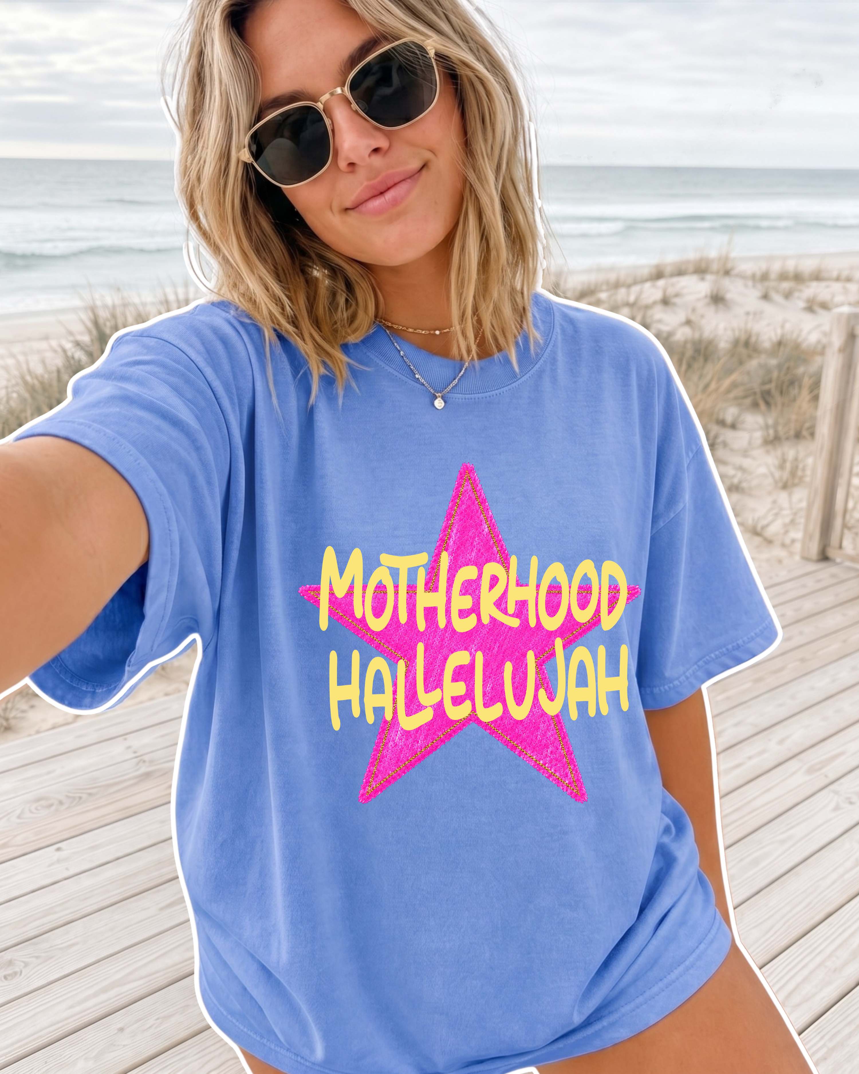 Woman taking a beach selfie wearing a flo blue Comfort Colors t-shirt with pink star graphic reading "Motherhood Hallelujah" in yellow lettering, ocean and dunes in background