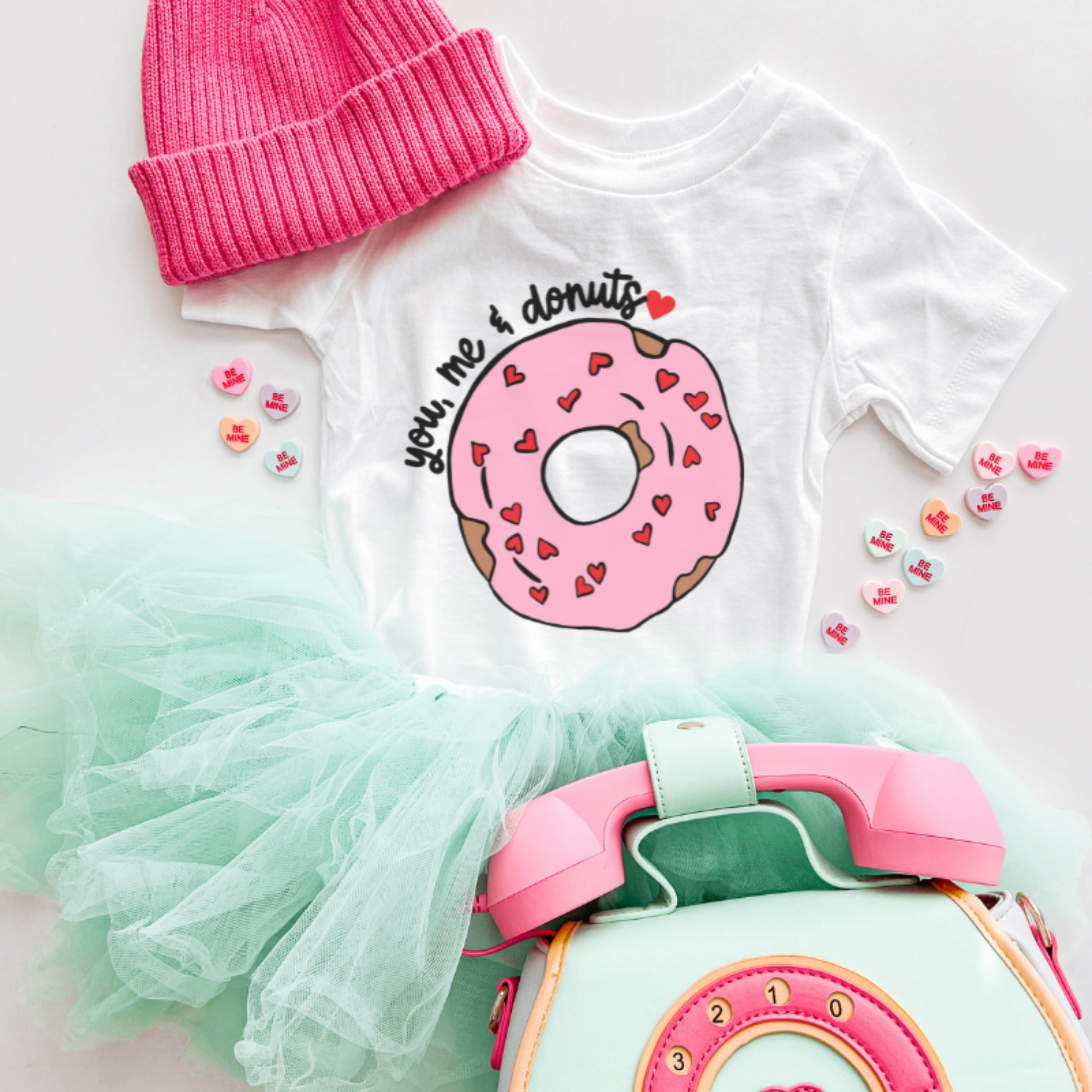 White t-shirt with pink donut graphic, pink beanie, green tutu, and pink toy on a white background